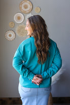 A woman with long wavy brown hair is shown from the back wearing a teal long-sleeve top and light blue jeans, standing against a gray wall with decorative woven wall art.