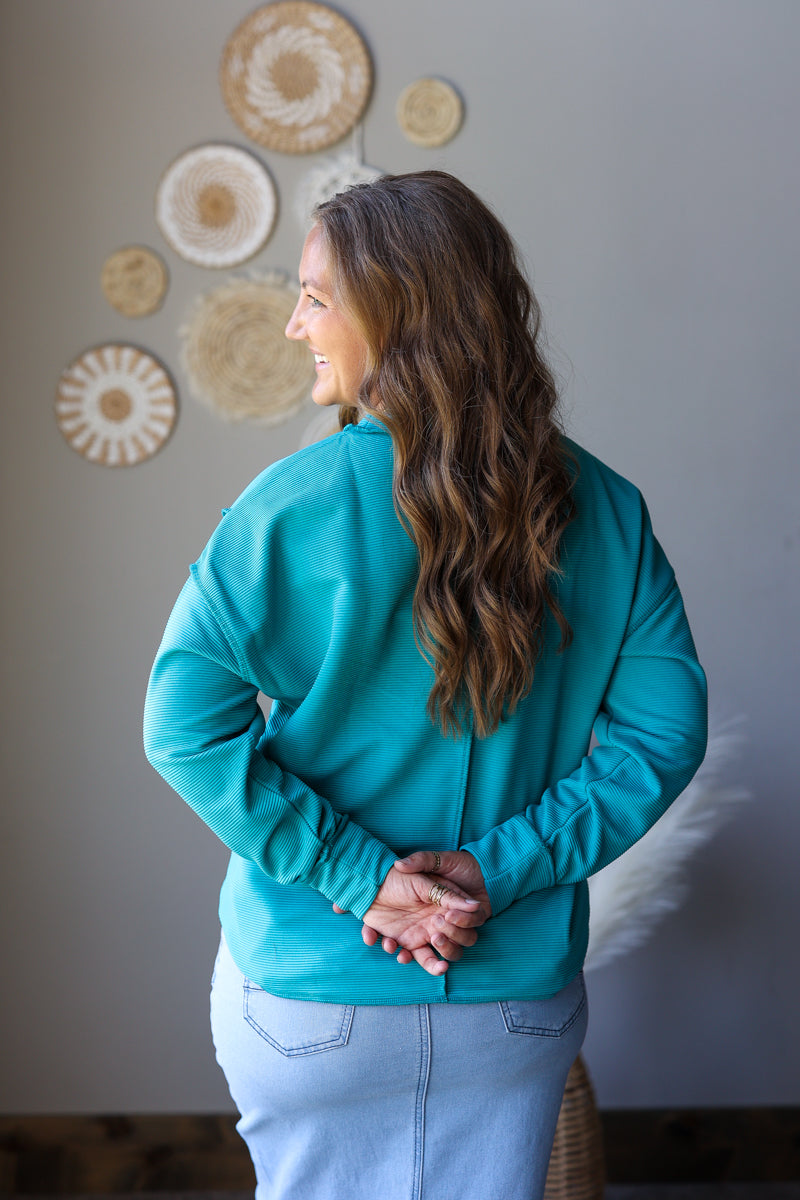 A woman with long wavy brown hair is shown from the back wearing a teal long-sleeve top and light blue jeans, standing against a gray wall with decorative woven wall art.