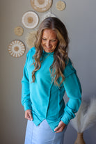 A woman wearing a bright turquoise long-sleeve top and light blue denim skirt, standing in front of a gray wall decorated with circular woven wall art.