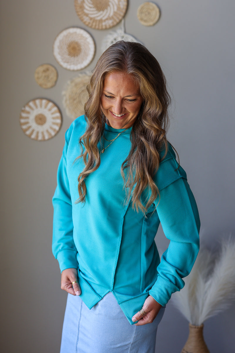 A woman wearing a bright turquoise long-sleeve top and light blue denim skirt, standing in front of a gray wall decorated with circular woven wall art.