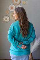 A woman with long wavy brown hair is shown from the back wearing a teal long-sleeve sweatshirt and light blue denim skirt, standing in a room with neutral-toned wall decor.