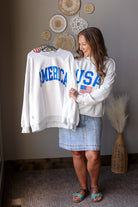 Woman holding a white sweatshirt with 'AMERICA' and 'USA' text, standing in a room with decorative items on the wall.