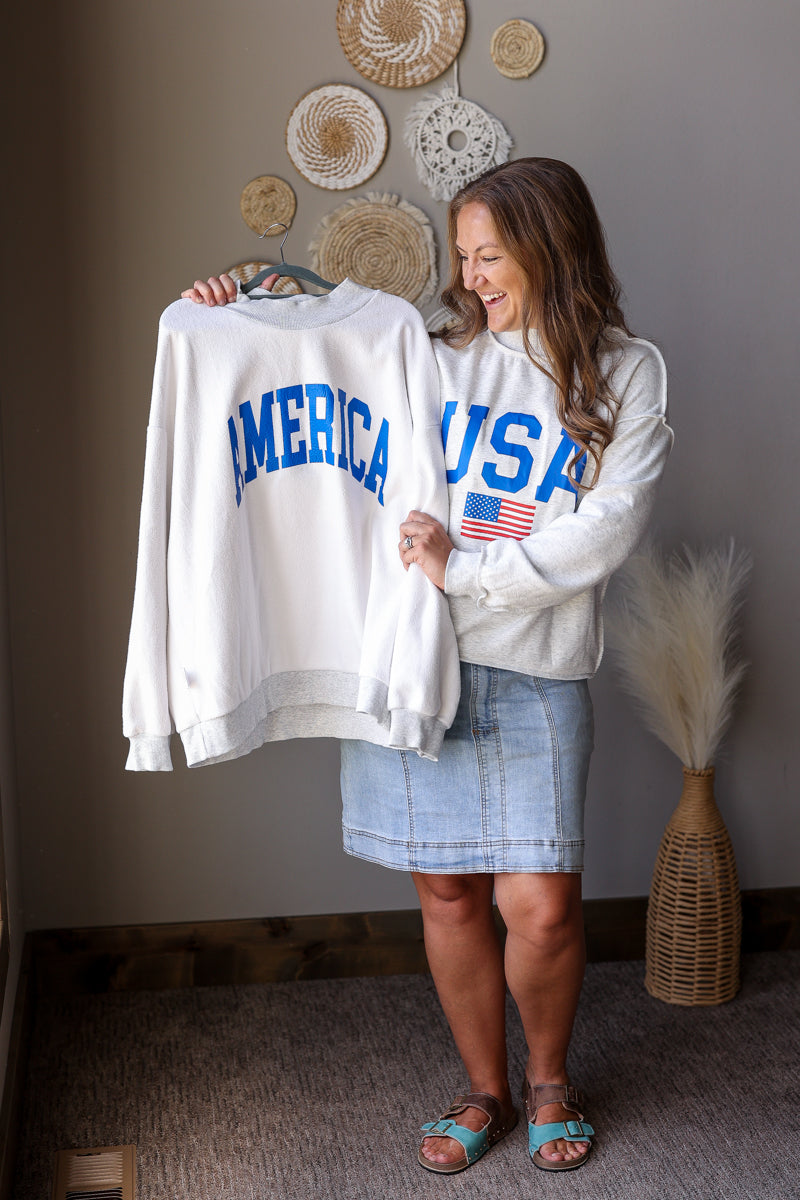 Woman holding a white sweatshirt with 'AMERICA' and 'USA' text, standing in a room with decorative items on the wall.