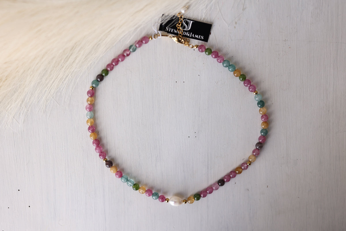 A beaded choker necklace with multicolored faceted beads and a small white pearl, displayed on a white wooden surface with a black brand tag attached.