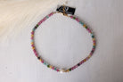 A beaded choker necklace with multicolored faceted beads and a small white pearl, displayed on a white wooden surface with a black brand tag attached.