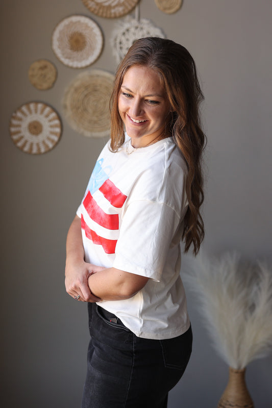 Woman wearing a white t-shirt with a red graphic design, standing against a gray wall with decorative items.