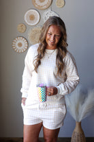 A woman wearing a matching cream and white gingham sweatshirt and shorts set, holding a colorful patterned tumbler, standing in a styled room with wall decor.