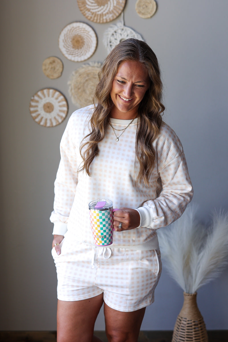A woman wearing a matching cream and white gingham sweatshirt and shorts set, holding a colorful patterned tumbler, standing in a styled room with wall decor.