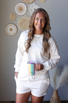 A smiling woman wearing a matching cream and white gingham lounge set, consisting of a long-sleeve top and shorts, holding a colorful patterned tumbler against a gray wall decorated with woven wall art.