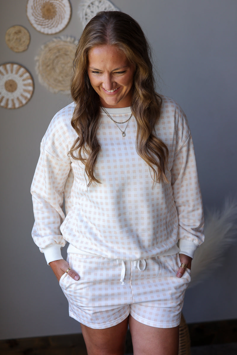 A woman wearing a matching two-piece lounge set with a white and beige gingham pattern, featuring a long-sleeve sweatshirt and shorts with a drawstring waist, standing against a gray wall with decorative wall art.