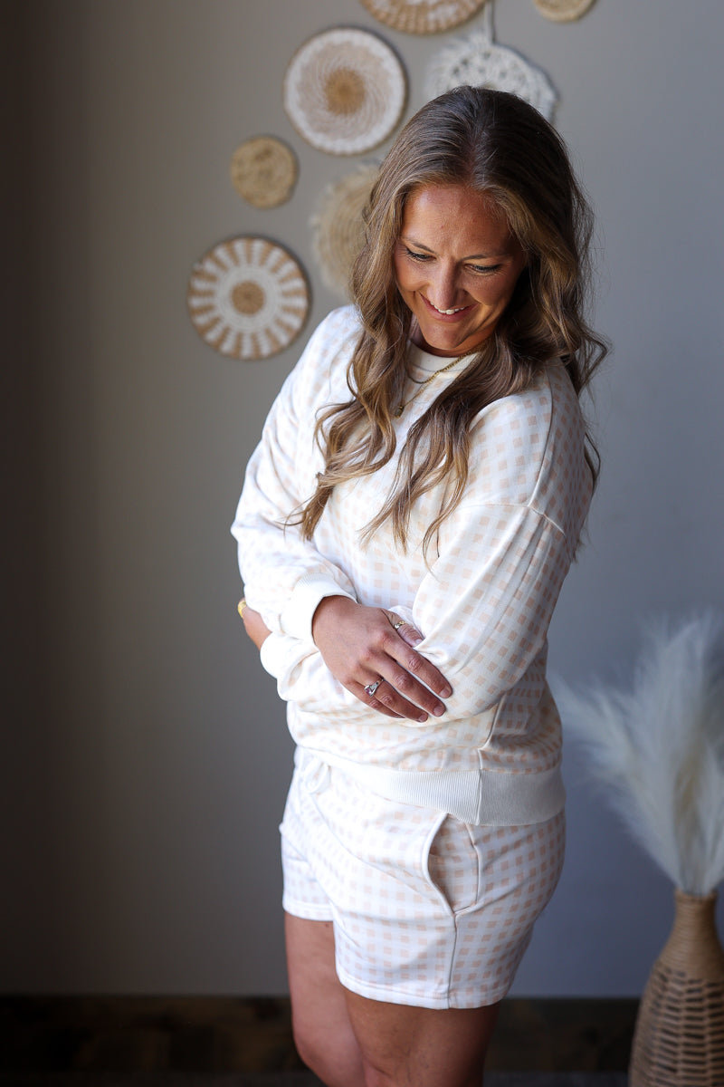 A woman wearing a matching white and beige gingham pajama set, consisting of a long-sleeve top and shorts, standing in a room with neutral-toned walls and decorative wall art.