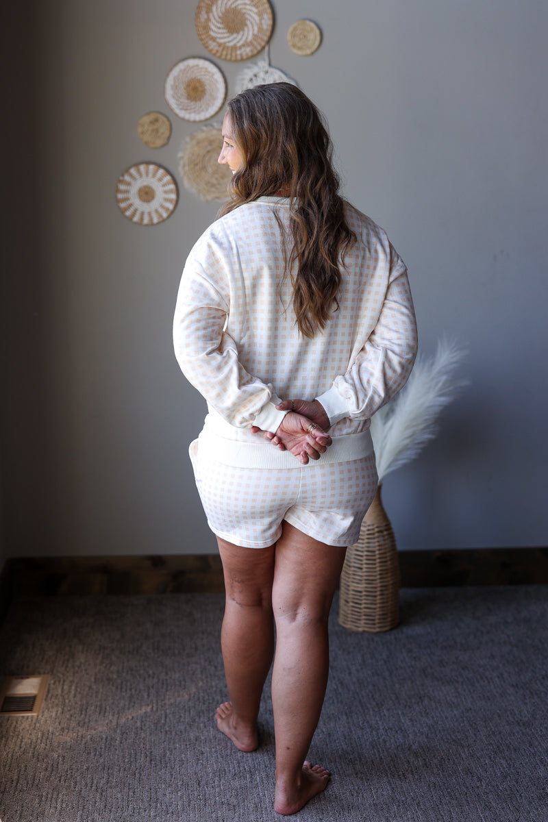 A woman stands with her back to the camera wearing a cream and white gingham long-sleeve top and matching shorts set, in a styled room with neutral decor.