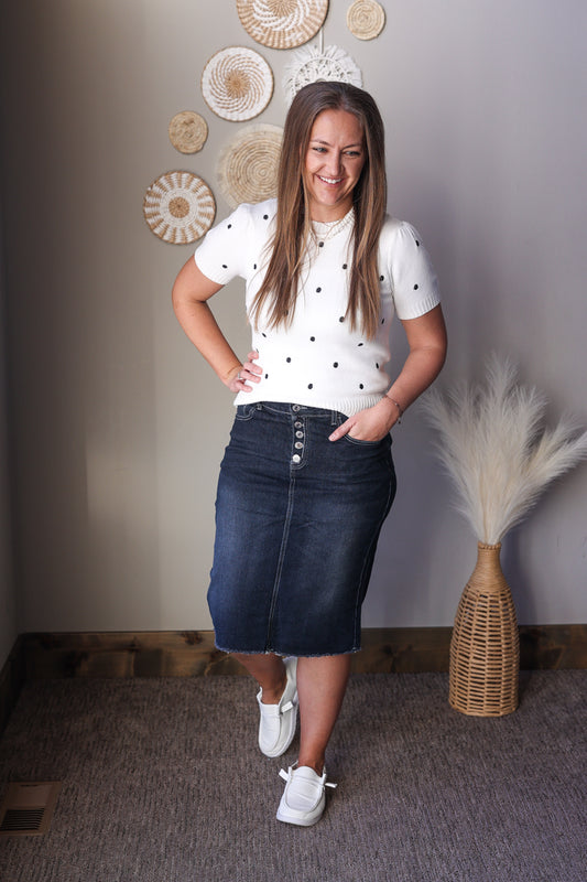 Woman wearing a white polka dot shirt and blue denim skirt standing in a room with decorative wall art and plants.