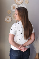 Woman wearing a white sweater with black polka dots and dark denim skirt, standing against a neutral wall with decorative elements.