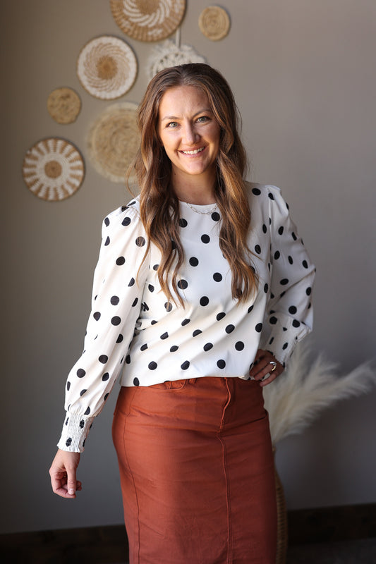 Woman wearing a polka dot blouse and brown pants standing against a wall with decorative items.