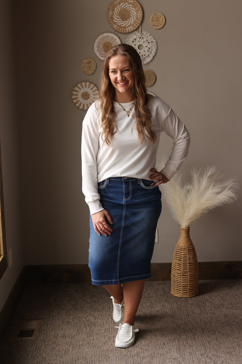 Woman wearing a white long-sleeve top and blue denim skirt standing in a room with decorative wall plates and a vase.
