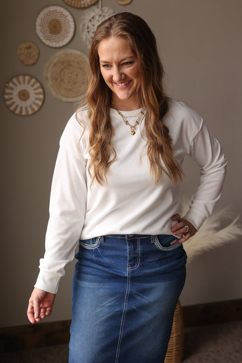 Woman wearing a white long-sleeve shirt and blue jean skirt standing against a wall with decorative plates.