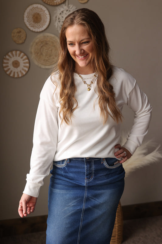 Woman wearing a white long-sleeve shirt and blue jean skirt standing against a wall with decorative plates.