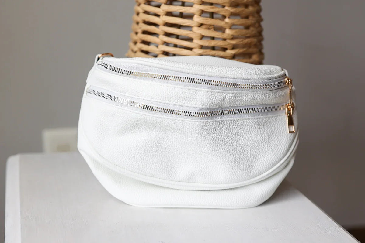 White leather waist bag with gold zippers on a white surface with a wicker basket in the background.