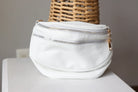 White leather waist bag with gold zippers on a white surface with a wicker basket in the background.