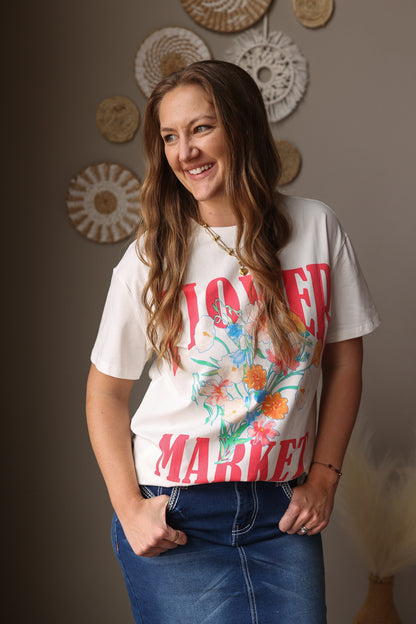 Woman wearing a white t-shirt with colorful graphics and text, standing against a neutral wall with decorative items.