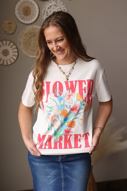 Woman wearing a white t-shirt with colorful floral design and text, standing against a neutral wall.