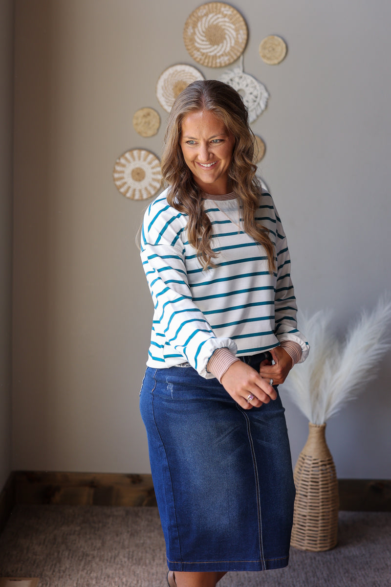 A woman wearing a long-sleeve white and teal striped knit top with a dark blue denim skirt, standing in a room with neutral-toned walls and decorative wall art.