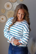 A woman wearing a white long-sleeve crewneck sweatshirt with teal blue horizontal stripes, paired with dark blue jean skirt, standing against a gray wall decorated with circular woven wall art.