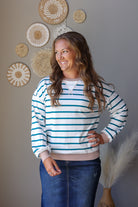 A woman wearing a white and teal blue striped long-sleeve crewneck sweatshirt with a ribbed beige hem, paired with a white undershirt and blue jean skirt, standing against a gray wall decorated with woven wall art.
