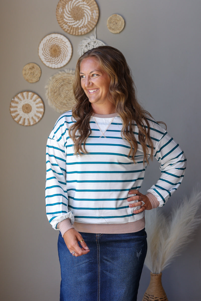 A woman wearing a white and teal blue striped long-sleeve crewneck sweatshirt with a ribbed beige hem, paired with a white undershirt and blue jean skirt, standing against a gray wall decorated with woven wall art.
