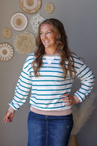 A woman wearing a white long-sleeve crewneck sweater with teal blue horizontal stripes and a ribbed pinkish-beige hem, paired with a white undershirt and blue jean skirt, standing against a gray wall decorated with woven circular wall art.
