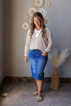 A woman modeling a cream and brown striped long-sleeve knit top with a white collar detail, paired with a blue denim midi skirt and teal sandals, standing in a room with a gray wall decorated with circular wall art.
