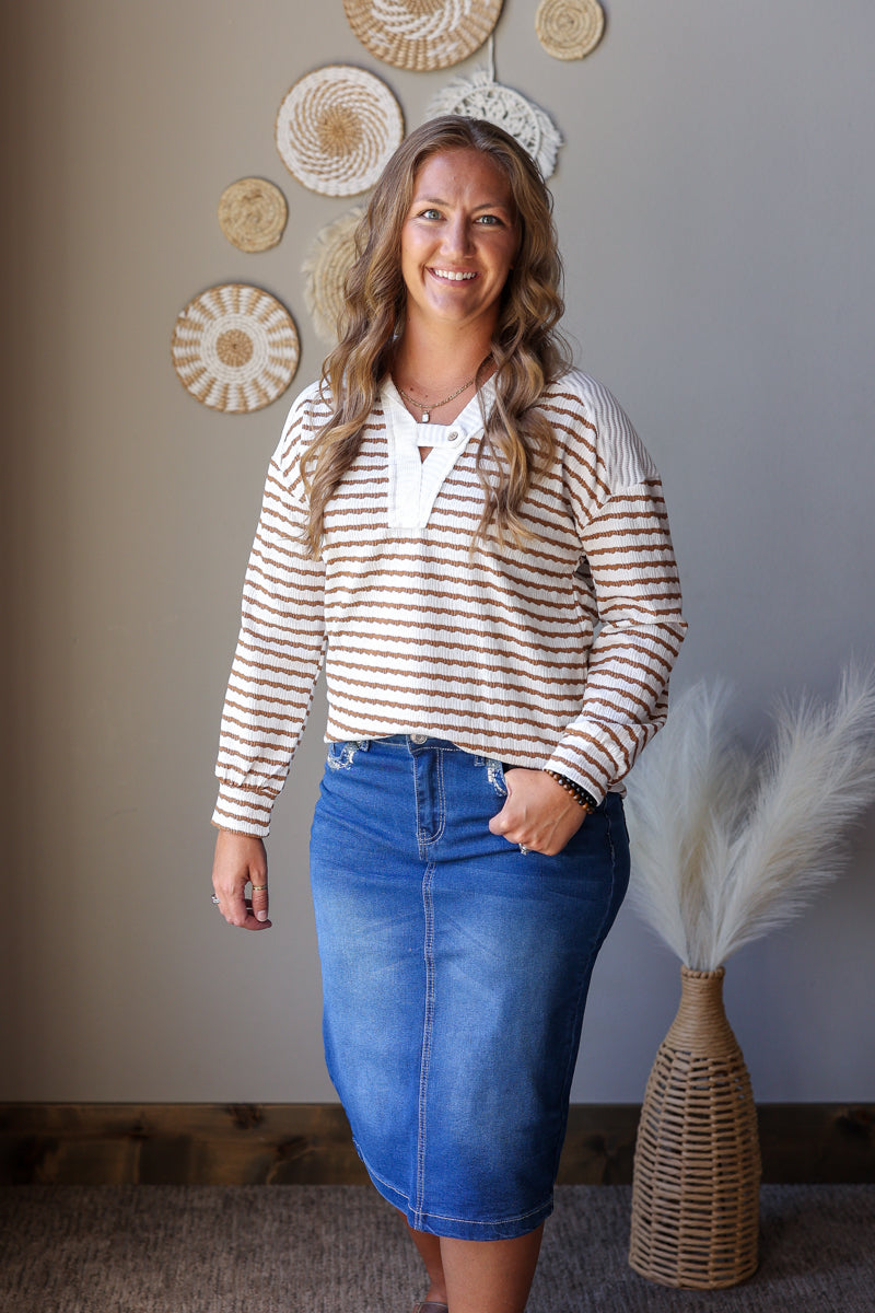 A woman modeling a cream and brown horizontally striped long-sleeve knit top with a white V-neckline, paired with a blue denim skirt, standing in front of a gray wall with decorative woven wall art.