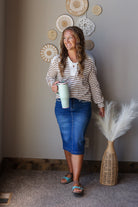 A woman stands indoors wearing a beige and brown striped long-sleeve top, a blue denim pencil skirt, and blue sandals. She holds a mint green travel mug.