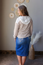 A woman is shown from behind wearing a cream and brown striped hoodie with a white knit hood, paired with a blue denim skirt, standing in a room with a neutral gray wall and decorative wall art.