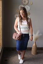Woman standing indoors wearing a striped tank top and blue skirt, holding a brown purse.