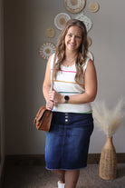 Woman standing indoors holding a brown leather bag, wearing a white sleeveless top with stripes and a blue denim skirt.