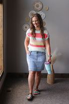 Woman wearing a colorful striped shirt and denim skirt holding a pink cup in a room with decorative wall art.