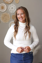 A woman with long wavy brown hair smiling, wearing a fitted white long-sleeve mock neck top and blue jean skirt, standing against a neutral gray wall decorated with bohemian woven wall hangings.