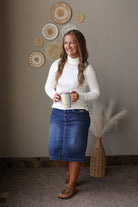 A woman stands indoors wearing a white mock-neck turtleneck sweater and a blue denim skirt, holding a patterned tumbler. She is posed in front of a neutral wall decorated with circular woven wall art and a vase of pampas grass.