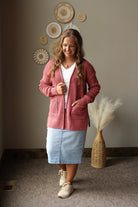 A woman modeling a dusty rose waffle-knit zip-up cardigan over a white t-shirt and a light blue denim skirt, standing in a styled room with decorative wall art and a vase.