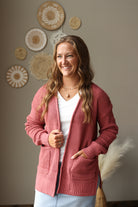 A woman wearing a dusty rose waffle-knit cardigan over a white V-neck top and light blue jean skirt, standing in a room with boho wall decor.