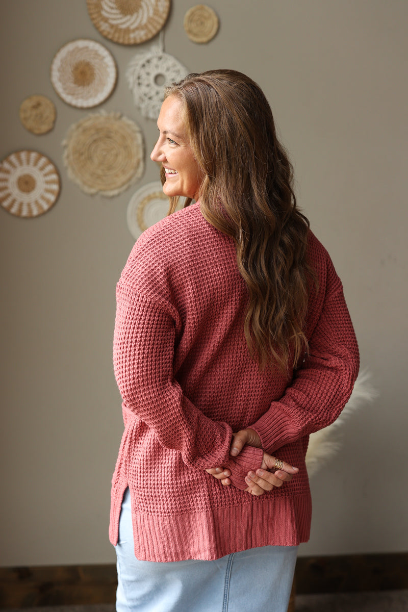 A woman with long brown hair is shown from the back wearing a dusty rose waffle-knit sweater and light blue jean skirt, standing in front of a neutral wall decorated with circular woven wall art.