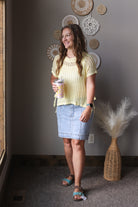 Woman in a yellow top and denim skirt holding a coffee cup, standing in a room with decorative wall art and a vase.