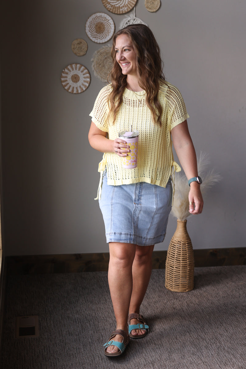 Woman in yellow top and blue skirt holding a cup, standing in a room with decorative wall art.
