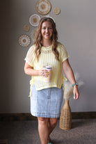 Woman in a yellow top and denim skirt holding a cup, standing in a room with decorative wall art.