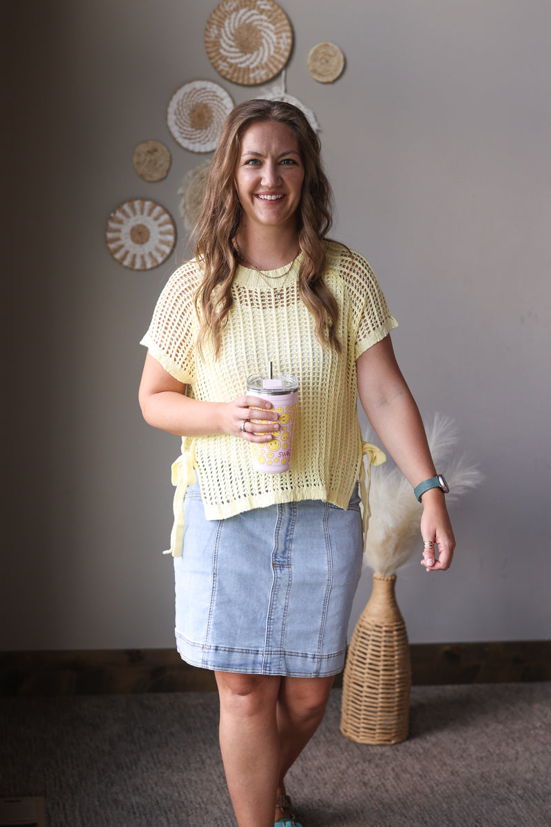 Woman in a yellow top and denim skirt holding a cup, standing in a room with decorative wall art.