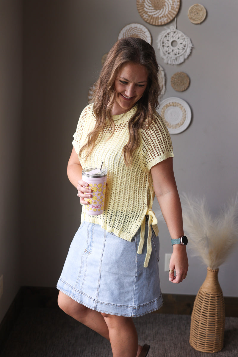 Woman in a yellow top and denim skirt holding a drink, standing in a room with decorative plates on the wall.