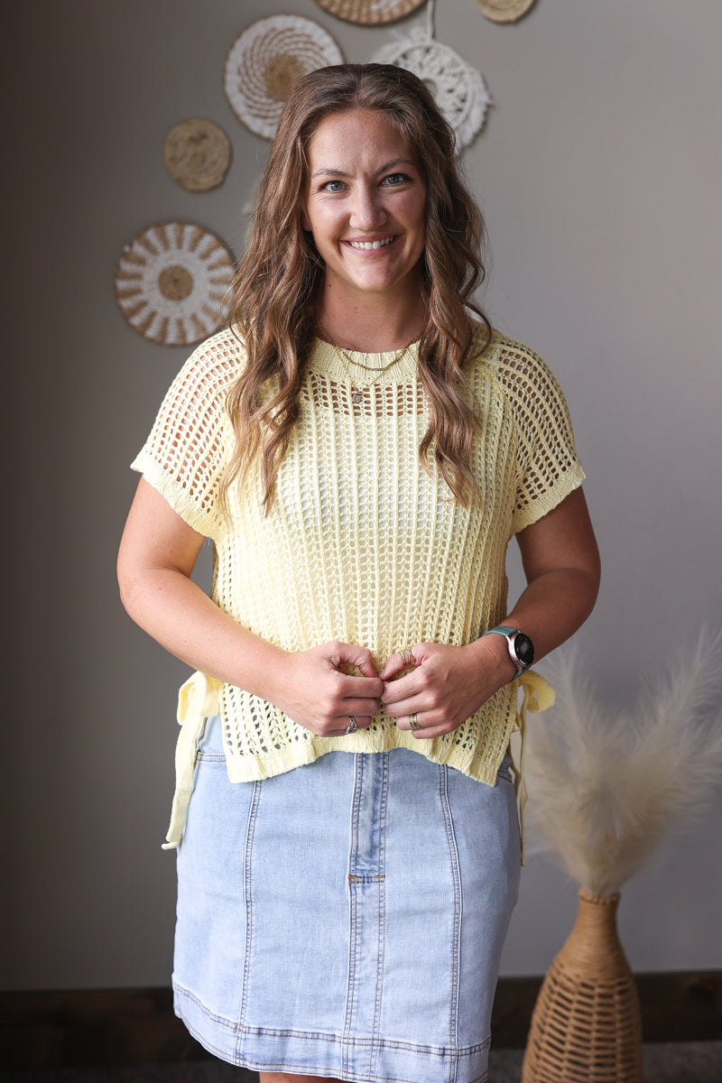 Woman wearing a yellow crochet top and denim skirt standing against a neutral wall with decorative elements.