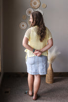 Woman wearing a yellow knitted sweater and denim skirt standing in a room with decorative wall items.